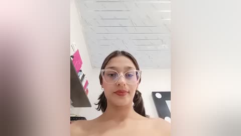 Video of a young woman with medium skin tone, dark hair, and glasses, standing topless in a brightly lit room with a white ceiling and pink and black artwork.