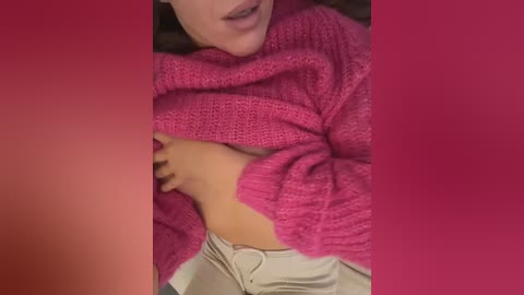 Video of a woman in a pink knitted sweater, partially lifting it to reveal her small, light-skinned breasts. She wears beige pants and has her lips slightly parted. Background is blurred.
