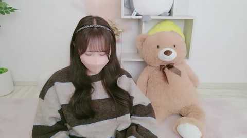 A young woman with long brown hair, wearing a striped sweater, sits on a light-colored rug. She is flanked by a large stuffed bear with a yellow hat. The background features white walls and a potted plant.