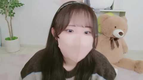Media: Video of a young Asian woman with long brown hair, wearing a white mask and a dark sweater, indoors, with a large brown teddy bear and a potted plant in the background.