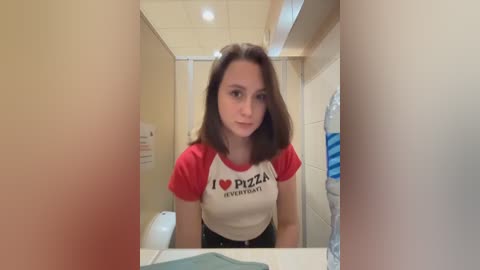 Media: A video of a young woman with straight brown hair, wearing a red and white \"I \u2665 PIZZA\" t-shirt, standing in a bathroom with beige tiles and a blue towel.