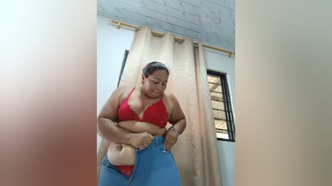 Media: A video of a plus-sized Latina woman in a red bra and blue jeans, adjusting her pants, standing in a room with beige curtains and a window.
