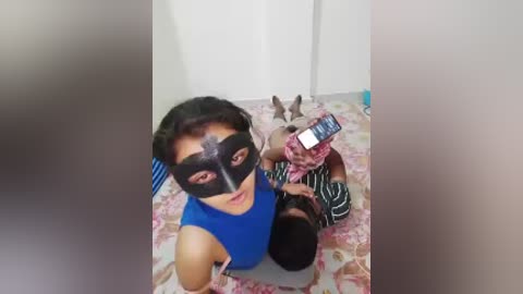 Video of a young woman with short dark hair and a black mask, sitting on a floral-patterned bed with a dog and a phone, in a plain, white-walled room.