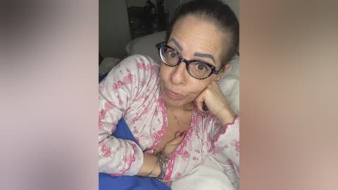 Video of a middle-aged woman with light skin, wearing glasses, and a pink tie-dye shirt, lying on a bed with white sheets and a blue blanket.