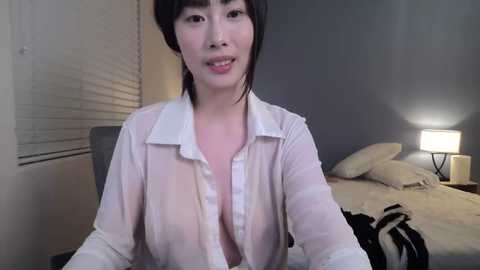 A video of an Asian woman with short black hair, wearing a sheer white blouse, standing in a dimly lit bedroom with a bed and a lamp.