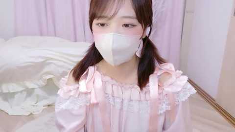 Video of an East Asian girl with straight brown hair in pigtails, wearing a light pink, frilly off-shoulder dress with lace trim, a white face mask, and standing in a room with white and pastel-colored furniture.