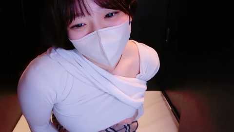 Video of an Asian woman with dark hair and a white mask, wearing a white long-sleeved top, partially covering her breasts, in a dimly lit room.