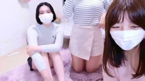 Video of three Asian women in casual attire, with face masks, sitting on a pink shag rug. One wears a striped sweater and white skirt, another a gray top and black shorts, and the third has a light sweater.