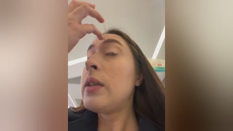 Video of a woman with light brown skin and long dark hair, eyes closed, being touched by another hand on her forehead, in a dimly lit room with white ceiling.