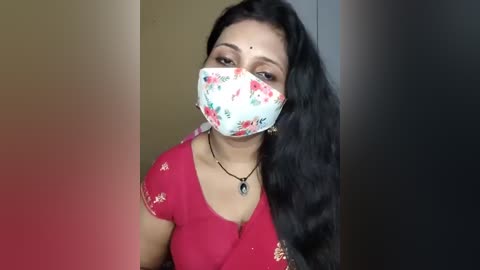 Video of an Indian woman with long black hair, wearing a red saree with floral embroidery, a floral face mask, and a black necklace. She stands against a blurred beige and dark background.
