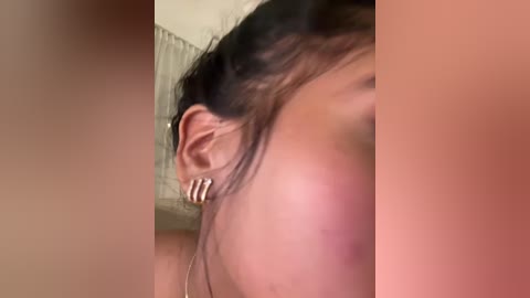 Media: A close-up video of a woman's ear with a silver hoop earring, showing her profile. The background is blurred, featuring a beige curtain.