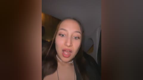 Media: Video of a young woman with light brown skin and long dark hair, wearing a black top, sticking her tongue out in a playful manner. The background is dimly lit with indistinct furniture.