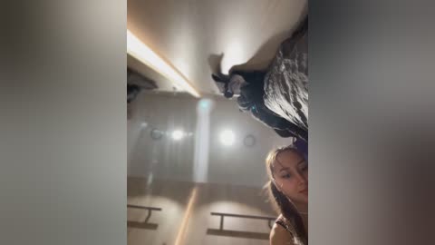 Media: Video of a young woman with light skin and brown hair partially obscured by a dark, tattered curtain hanging from the ceiling, with bright overhead lights illuminating the background.