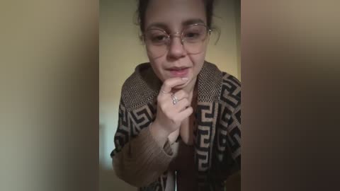 A video of a young woman with fair skin, glasses, and dark hair in a ponytail, wearing a brown and white geometric-patterned cardigan. She's indoors, looking contemplative with her finger to her lips.