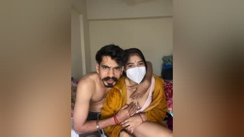 Video of a young couple, a man with short hair and a beard, and a woman with dark hair and a face mask, in a dimly lit room, wearing casual clothing.