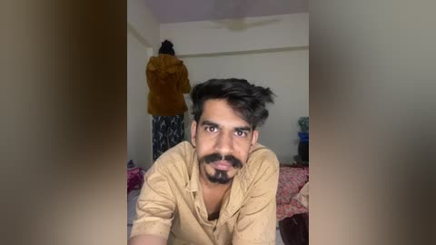 Video of a young man with medium brown skin, dark hair, and beard, wearing a beige shirt, lying on a bed in a simple room with a ceiling fan and a woman standing behind him.