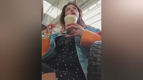 Video of a young woman with long brown hair, wearing a black dress with white polka dots, a blue denim jacket, and orange knitted sleeves. She's holding a beige cup, sitting in a booth with brick walls and large windows in a cozy, casual setting.