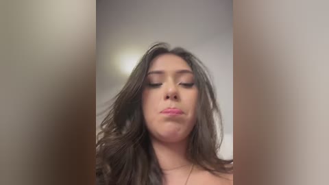 Video of a young woman with long, wavy dark hair, fair skin, and closed eyes, making a pouty face. Background is blurred with beige and brown tones.