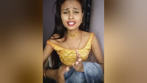 Media: A video of a South Asian woman with medium-brown skin, long black hair, and a gold choker, wearing a yellow off-shoulder blouse, and blue denim skirt, gesturing with her hands, in a dimly lit room.