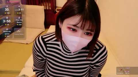 A video of an Asian woman with short black hair, wearing a white face mask and black-and-white striped top, sitting on a tatami mat in a traditional Japanese room.