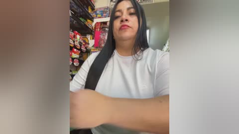 Video of a Latina woman with long black hair and medium skin tone, wearing a white long-sleeve shirt, shopping in a brightly lit convenience store filled with colorful snacks and beverages.