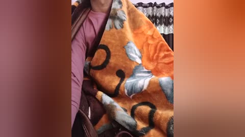Media: Video of a person sitting on a couch, wrapped in a vibrant orange blanket with a black and blue pattern, wearing a purple long-sleeved shirt. The background shows a partially visible wardrobe.