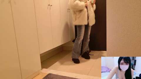Media: A video of a person in a beige fur coat, blue jeans, and black shoes, standing in a narrow hallway with white cabinets and a beige tiled floor. Inset: a woman with black hair and a black top.