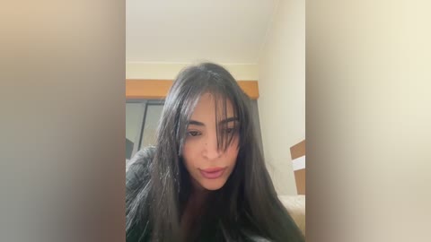 Media: A video of a young woman with long, straight black hair, wearing a dark green sweater, in a well-lit, beige-walled room with a wooden door and a white bed.