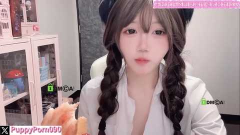 A video of a young Asian woman with long, dark brown hair in twin braids, wearing a white blouse. She holds a pink and white cake. Background features a shelf with food products.