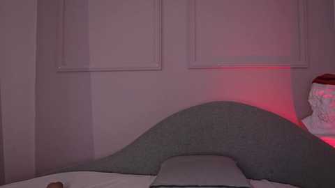 Video of a bedroom corner with a soft, curved headboard, gray bedding, and a red light casting a warm glow on the walls and headboard, giving the room a cozy, intimate atmosphere.