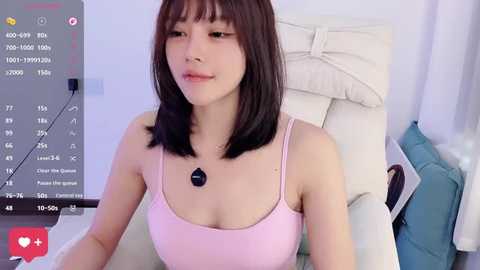 Video of a young East Asian woman with shoulder-length dark hair and light skin, wearing a pink spaghetti-strap tank top, sitting on a white couch, with a smartphone showing live stream icons in the background.