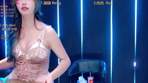 A video of a woman with fair skin, wearing a light pink, lace-trimmed, low-cut top, standing in a dimly-lit room with blue LED lighting.