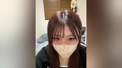 Video of an Asian woman with long, straight brown hair, wearing a beige mask and black cardigan, sitting in a bedroom with a bed, blinds, and stuffed animal.