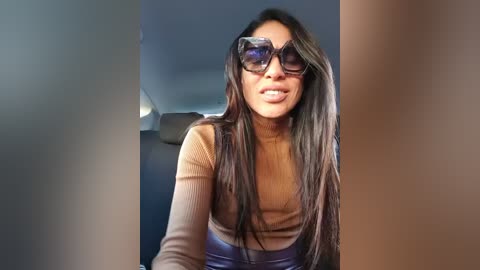 A video of a smiling woman with long, straight dark hair, wearing large, dark sunglasses, a mustard-yellow ribbed top, and blue jeans, standing indoors against a plain, light-colored wall.