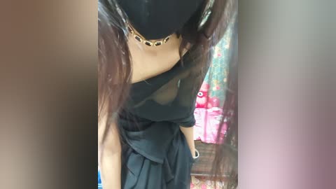 Video of a woman with long, dark hair wearing a black, sheer one-shoulder top and pleated skirt, standing in a brightly lit room with colorful toys in the background.