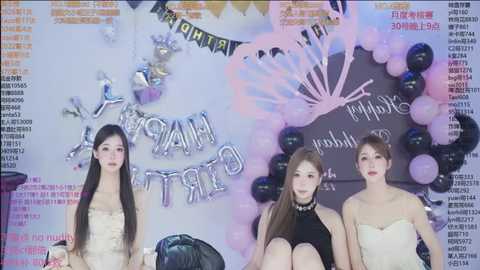 Video of three young Asian women with long hair, seated at a party with pastel balloons and \"Happy Birthday\" banners. They wear different outfits, one in a strapless dress, another in a black top, and the third in a beige top.