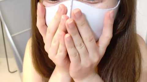 Video of a person with long, straight brown hair, wearing a white surgical mask, holding it over their mouth and nose. The background is blurred, showing a white wall and a metal shelf.