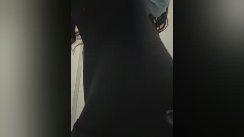 A dimly lit video of a person's upper body and arm, partially obscured by a dark background. The individual is wearing a light blue shirt and a dark jacket, with visible shadows suggesting a casual, indoor setting.