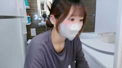 Media: Video of a young Asian woman with light skin and brown hair in a bun, wearing a gray shirt and white mask, sitting on a white bathtub edge in a modern bathroom with tiled walls and white sink.