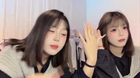 Video of two young Asian women with long black hair, wearing black clothing, smiling while one styles the other's hair. Background features white curtains and clothes hanging.