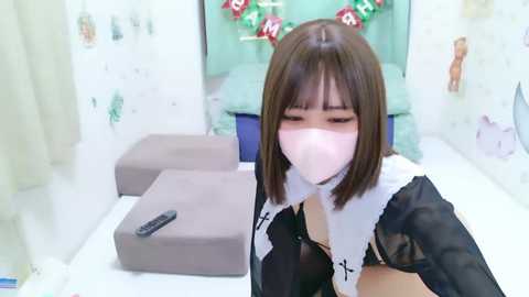 Media: Video of a young Asian woman in a maid outfit, wearing a pink mask, standing in a bright, clean room with a TV remote on a gray cushion.