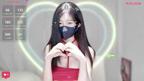 Video of an Asian woman with long black hair, wearing a red dress and blue mask, making heart shapes with her hands against a heart-shaped rainbow background.
