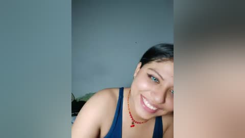 Video of a young woman with light brown skin and dark hair, smiling broadly. She wears a blue tank top and a red necklace. The background is a plain, light gray wall.