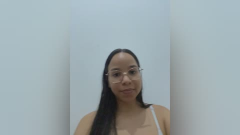 Video of a young woman with straight black hair, wearing glasses and a white tank top, standing against a plain white wall.