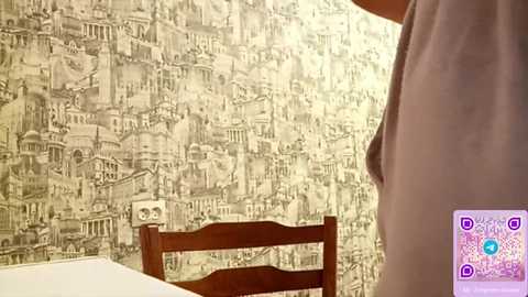 Video of a person in a light-colored tank top standing near a wooden chair with a beige patterned wallpaper depicting a cityscape in the background.