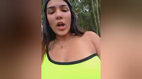 Media: Video of a young woman with medium brown skin, long dark hair, and closed eyes, wearing a neon green top with black trim, outdoors.