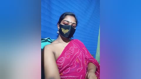 Video of a South Asian woman with medium skin tone, wearing a bright pink sari with gold embroidery, black face mask, and green saree draped over her shoulder. Background is a gradient of blue and purple, creating a vibrant, colorful scene.