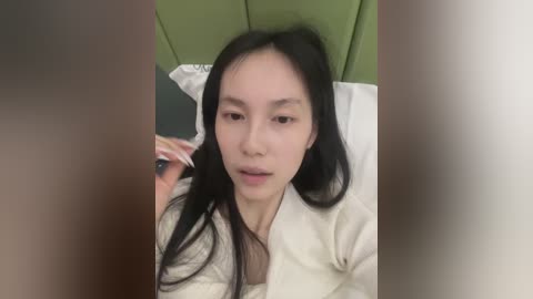 A video of an Asian woman with long black hair, wearing a white robe, lying on a green headboard, holding a phone, blurry background.