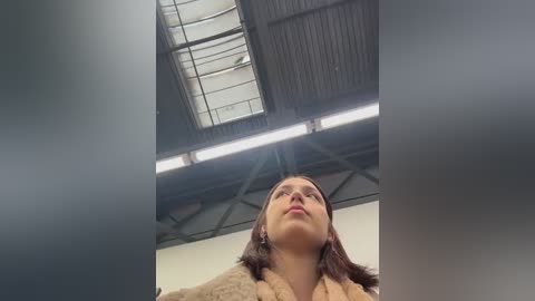 Video of a young woman with fair skin, straight brown hair, and a beige coat, looking upwards under a modern industrial ceiling with exposed beams and a large skylight.