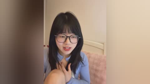 Video of a young Asian woman with shoulder-length black hair, wearing glasses, a light blue shirt, and light makeup, sitting on a pink couch in a beige room, looking at the camera with a slight smile.